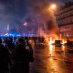 Vue nocturne d’émeutes à Paris avec fumée, barricades en feu et forces de l’ordre en intervention.